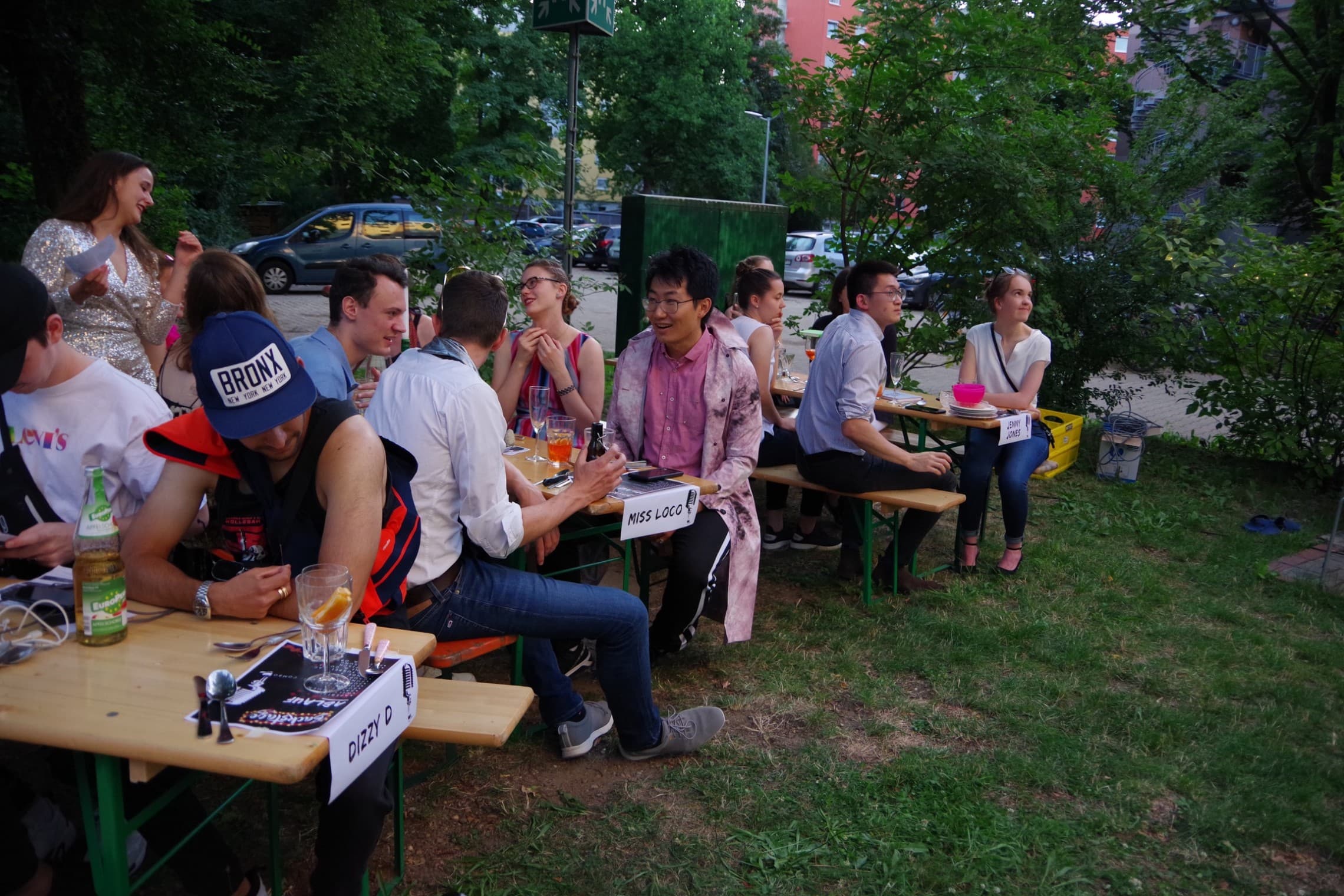 Ein Auschnitt des HEKdinners unter dem Motto 'Krimidinner'. Verkleidete Menschen sitzen an Bierbänken auf einer Wiese