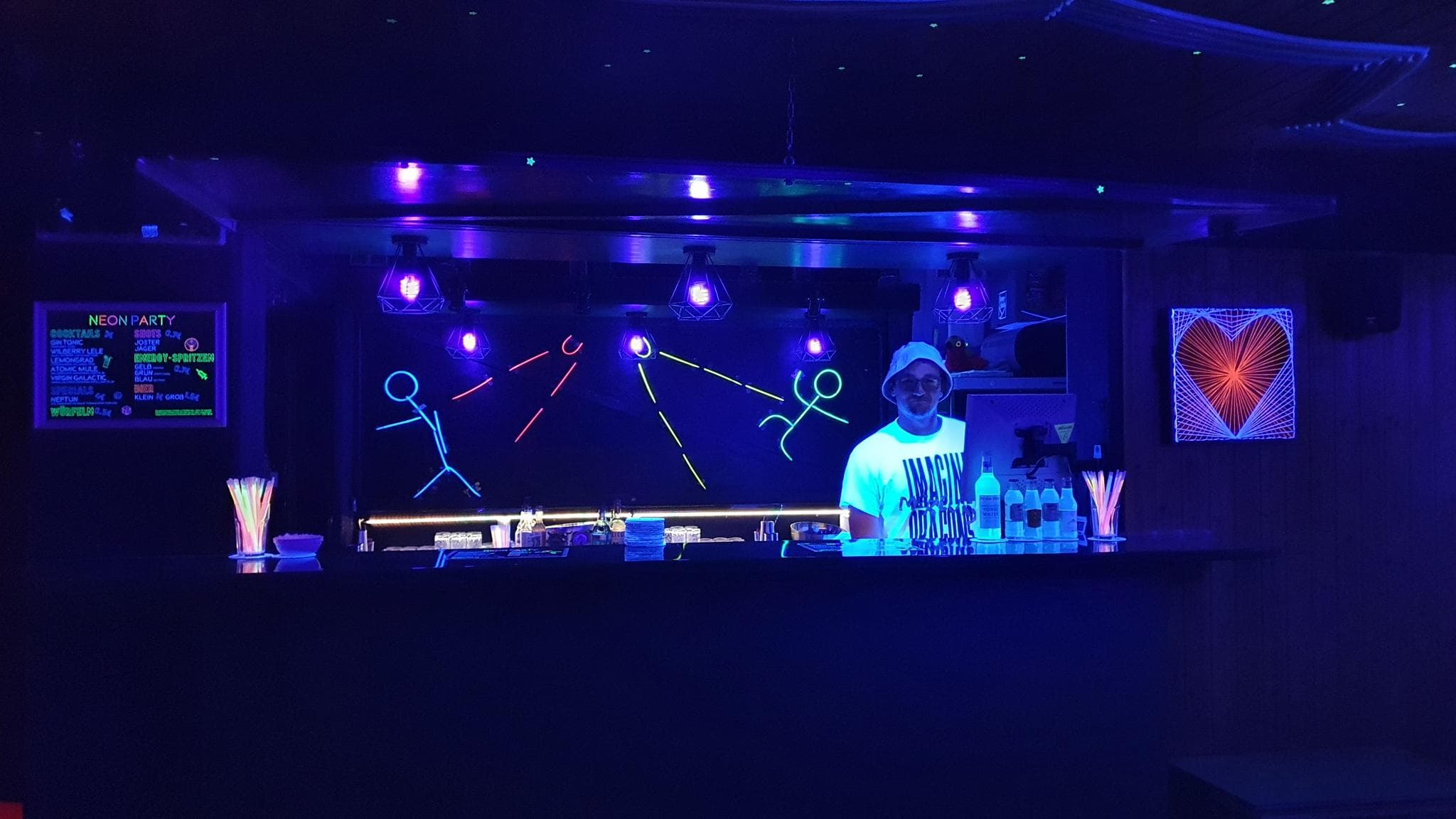 Wohnheimsbar dekoriert mit Schwarzlichtfarben für die Afterparty des Neueingezogenenabend. Ein Mensch steht hinter dem Tresen von einer mit Schwarzlichtfarben erleuchteten Bar.