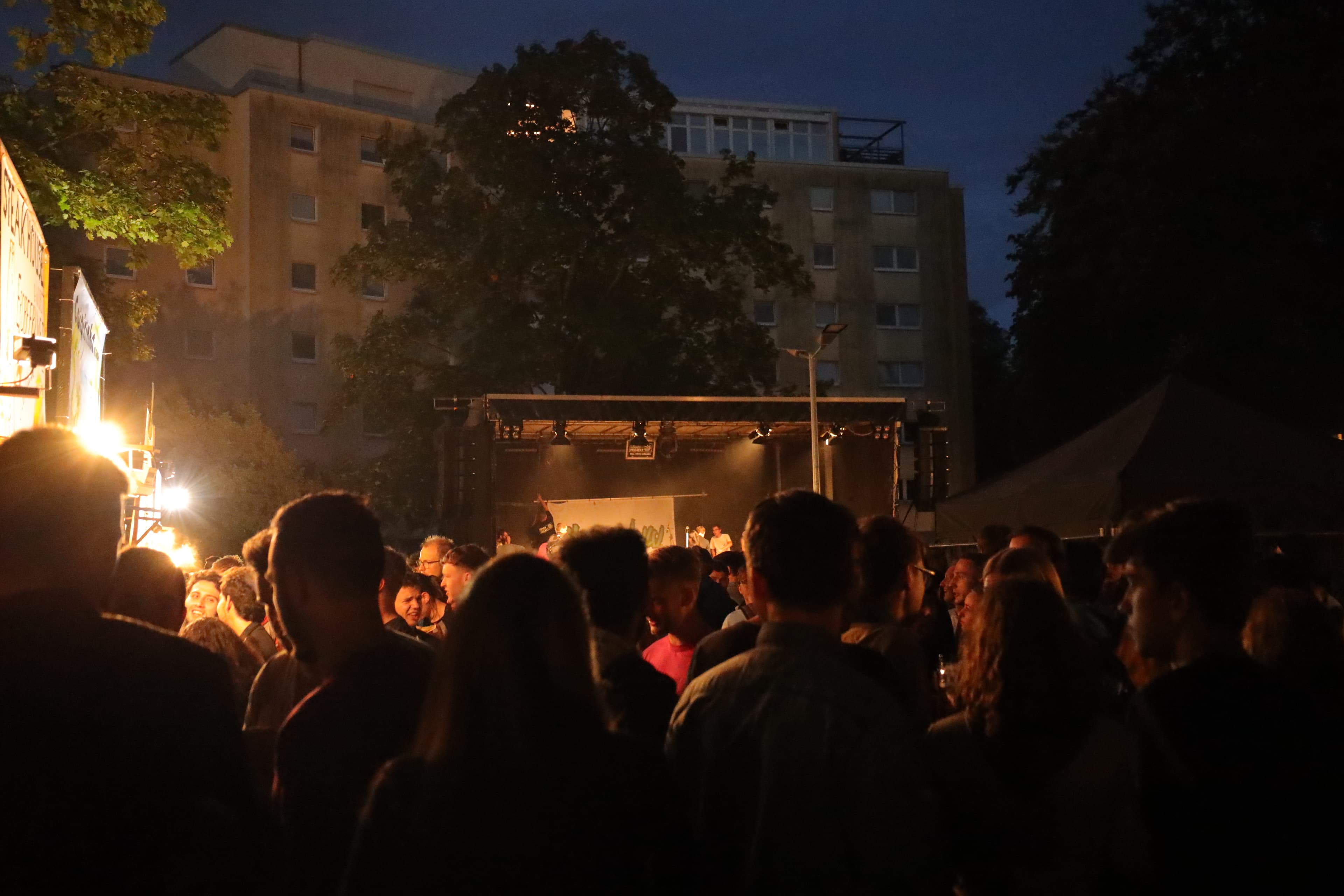 Feiernde Menschen vor einer Bühne beim Parkplatzfest mit HEK im Hintergrund. Feiernde Menschen vor einer Bühne beim Parkplatzfest mit Hermann-Ehlers-Kolleg im Hintergrund